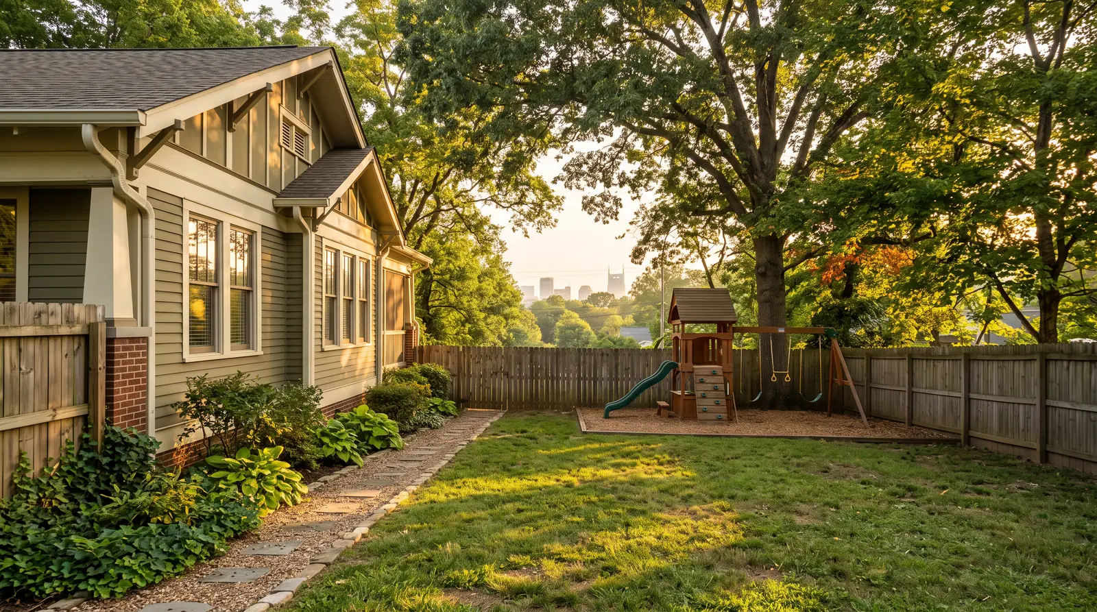 A charming East Nashville craftsman bungalow seen from the side yard at golden hour with a compact cedar playset and swing beam in the backyard, mature trees and downtown buildings faintly visible far behind