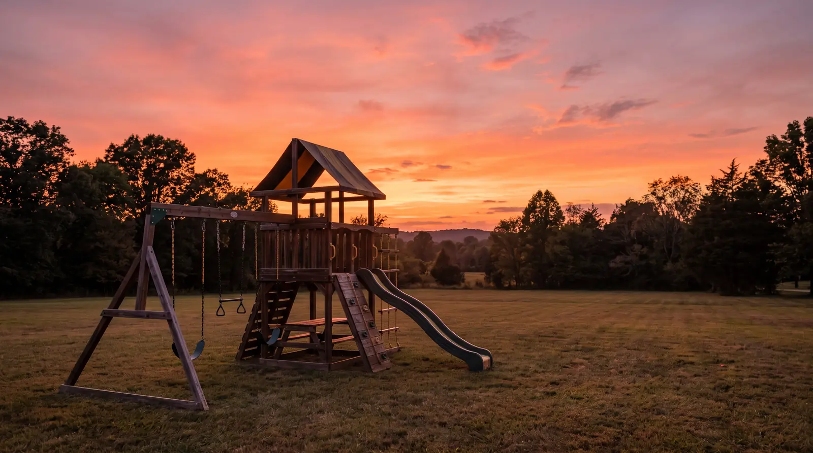 Playset Silhouetted by a Tennessee Sunset | Music City Playgrounds A cedar playset with a canopy roof and swings silhouetted against a glowing orange and pink Tennessee sunset sky, calm empty backyard